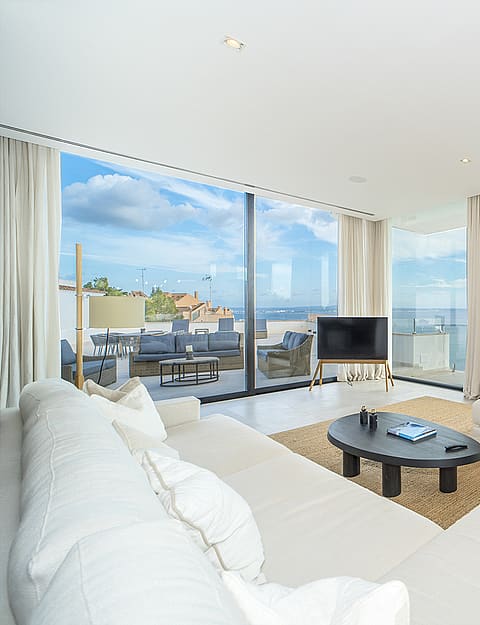 Modern living room featuring floor-to-ceiling glass walls, white sofas, woven area rug, and panoramic coastal views.