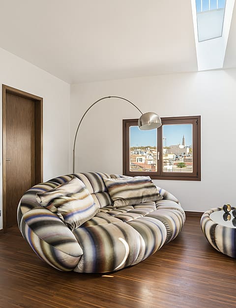 Modern living room with a striped round sofa, dark wood floors, a skylight, and a window viewing Palma Old Town.
