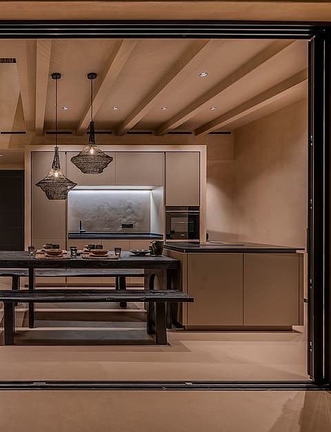 Interior view from a terrace looking into a minimalist kitchen with a wooden dining table, bench seating, and soft recessed lighting.