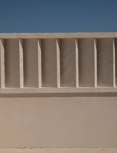 Close-up of a beige sandstone wall with rhythmic vertical recesses creating deep shadows under bright daylight.
