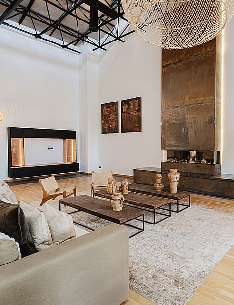 A spacious living area featuring a double-height ceiling with exposed black metal trusses, a tall rusted-metal fireplace surround, and woven pendant lighting.
