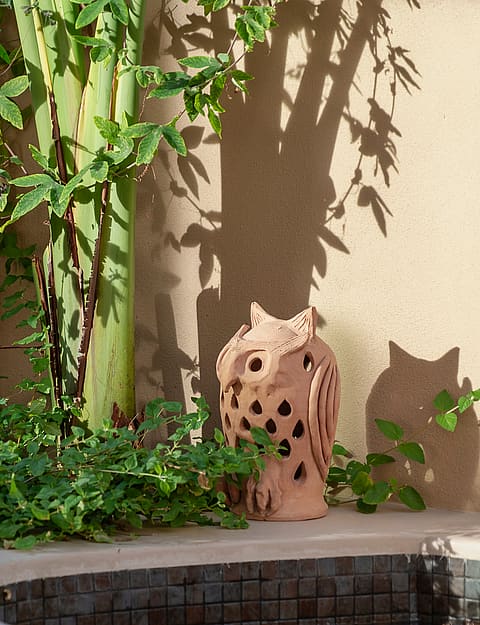 A terracotta owl lantern sits on a curved wall edge in a small garden corner with green foliage and textured beige wall.