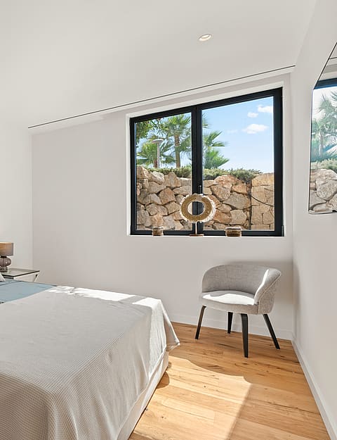 A minimalist bedroom featuring a grey upholstered bed, glass-topped side tables, and a view of a dry-stone wall outside.