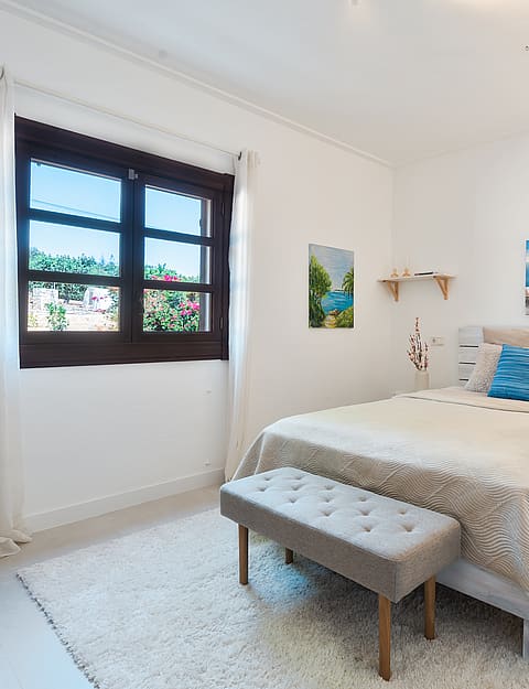 Dormitorio con cama de matrimonio, alfombra de texturas y marco de ventana de madera oscura hacia el verde.