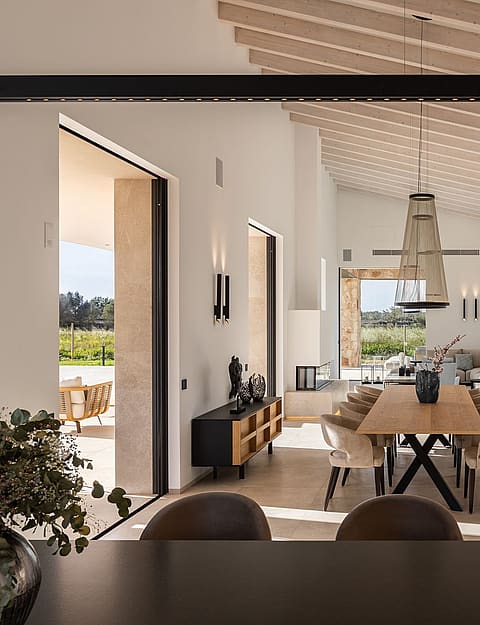 A spacious, bright open-plan room with white walls, wooden beamed ceilings, a large dining table, and direct terrace access.