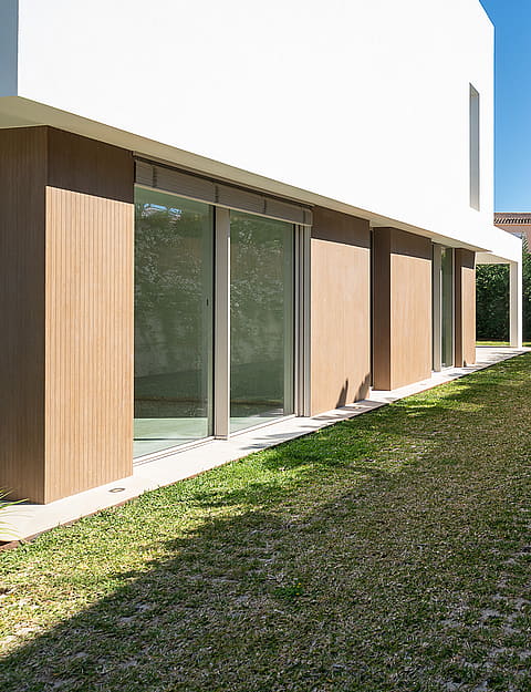 Side garden of a modern villa featuring a well-maintained lawn, white exterior wall with wood panels, and a blooming hedge.
