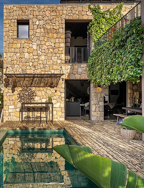 Exterior view of a stone-clad villa in Genova featuring a rectangular swimming pool, lush greenery, and climbing vines.