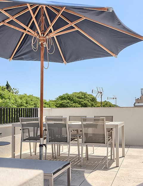Rooftop terrace with a dining table and chairs shaded by a dark blue umbrella, set against a bright sky.