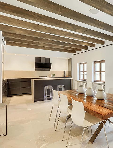 A bright open-plan kitchen and dining space with exposed wooden ceiling beams, a long wooden table, and minimalist white chairs.