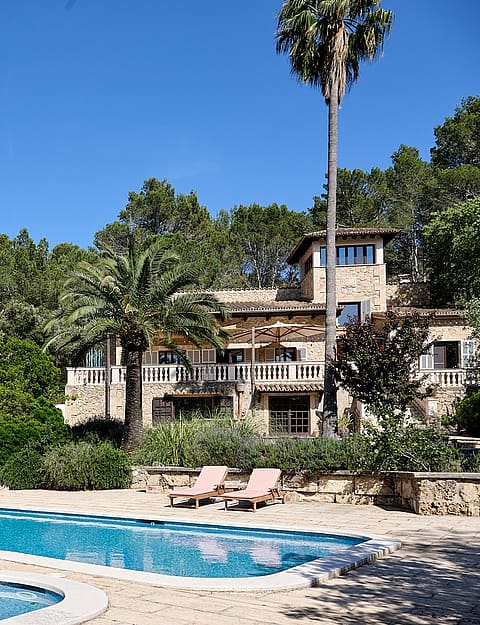 Exterior view of a stone finca with a large swimming pool, surrounding gardens, and mature palm trees under a clear sky.