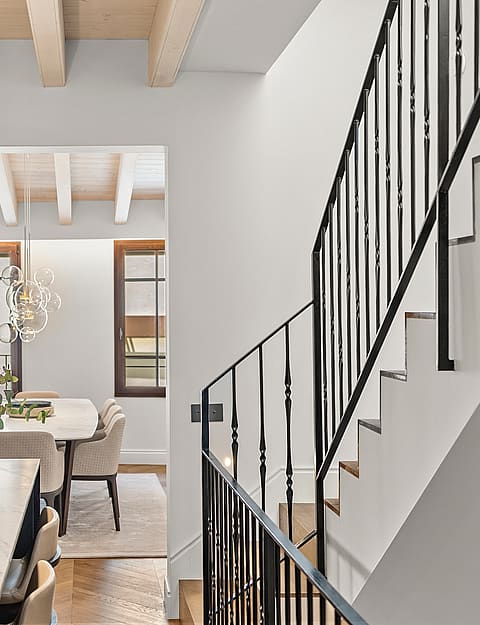 Interior view showing a black iron staircase railing, marble kitchen counter, and dining table under wooden ceiling beams.