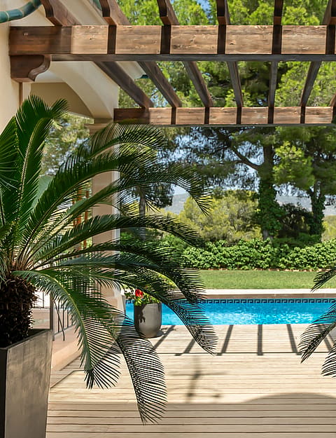 A wooden pergola shades a terrace overlooking a rectangular blue swimming pool with lush garden foliage in the foreground.