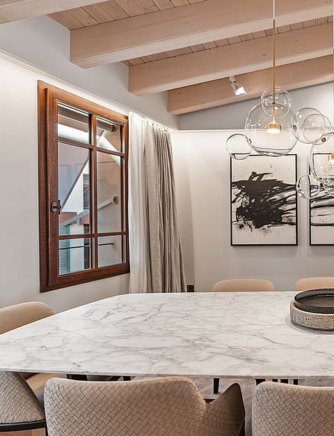 A modern dining space featuring a white marble table, glass globe pendant lights, and exposed light-wood ceiling beams.