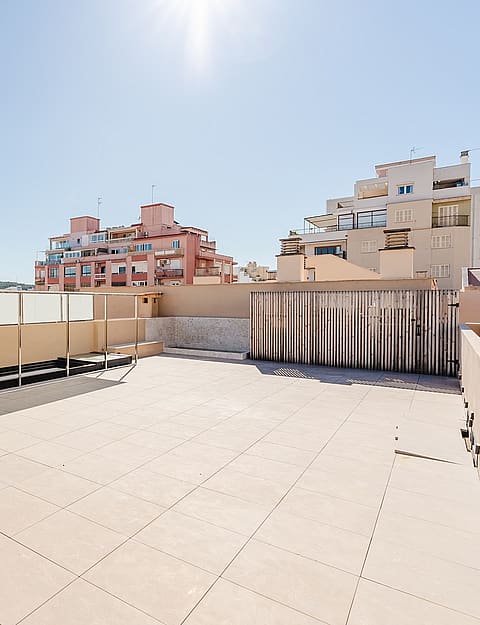 A wide, empty rooftop terrace with tiled flooring and low walls, overlooking surrounding city architecture under a clear blue sky.