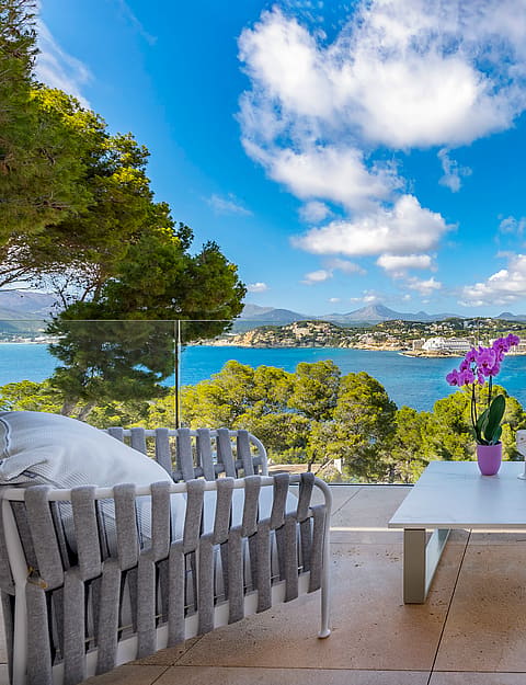 Modern outdoor terrace featuring woven grey furniture, a white coffee table with orchids, and a glass balustrade overlooking a bay with mountains in the background.