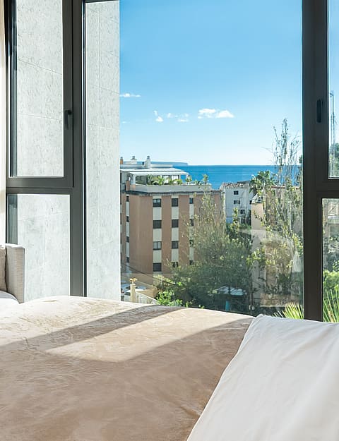 A partial view of a bedroom interior with a large window framing a sea view, featuring a tan bed cover and soft white curtains.