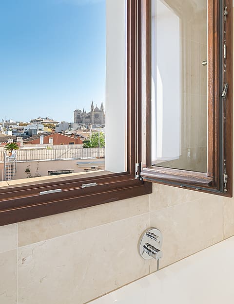 Modern bathroom interior featuring a white bathtub and glass shower next to an open window overlooking Palma's rooftops.