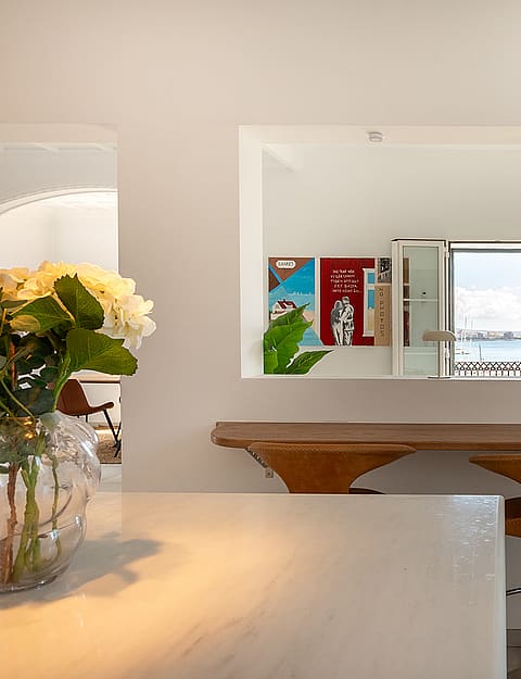 A white minimalist kitchen with a wooden bar stool and a rectangular pass-through window framing an ocean view.