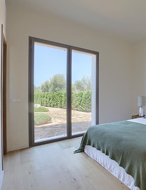 Bedroom with light wooden floors, a bed with a green spread, and a glass door opening to a terrace with garden views.