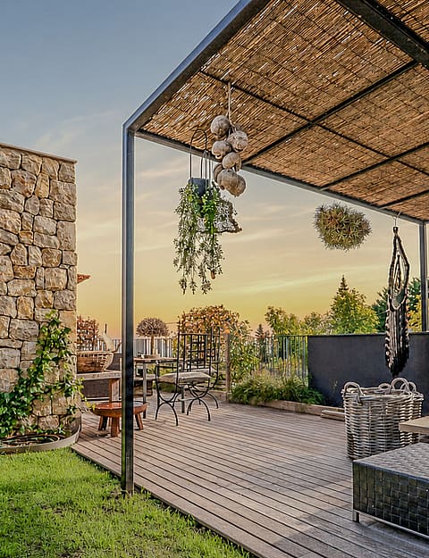 Covered outdoor terrace with wooden decking, stone wall, and metal pergola framing mountain views at sunset.