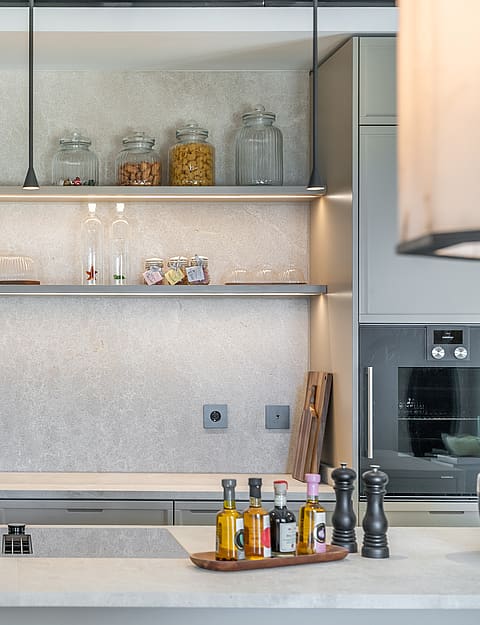 A contemporary kitchen showing a textured stone backsplash, slim floating shelves with LED lighting, and grey cabinetry.