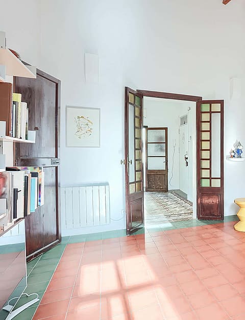 A bright living room with reddish terracotta tile floors, white walls, and a large sectional sofa. Wooden exposed ceiling beams are visible above. A set of open glass-paneled doors leads to an adjacent room.