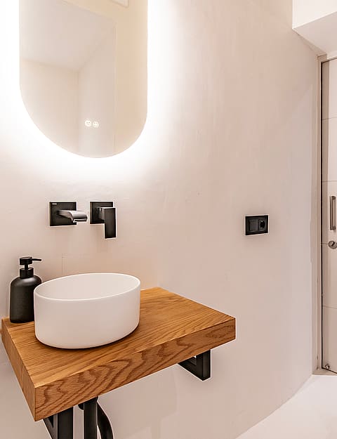 A modern guest bathroom featuring a wall-mounted wooden vanity, a white vessel sink, black wall-mounted faucet, and backlit mirror.