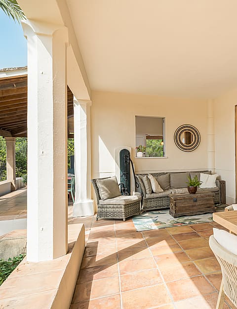 Covered outdoor terrace with terracotta tile flooring, wicker lounge seating, and a solid wood dining table in sunlight.