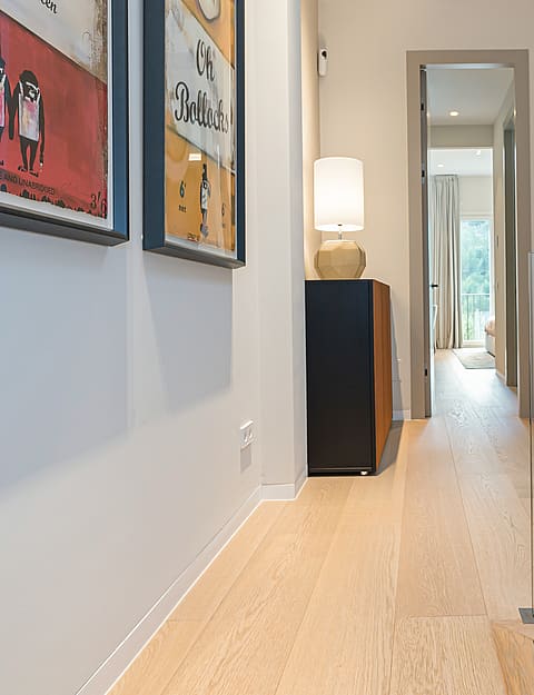 A bright residential hallway with light oak flooring, white walls, a glass stair railing, and a small sitting area.
