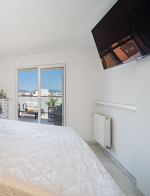 A bright bedroom with a queen bed, upholstered beige headboard, and a glass sliding door leading to an outdoor balcony.