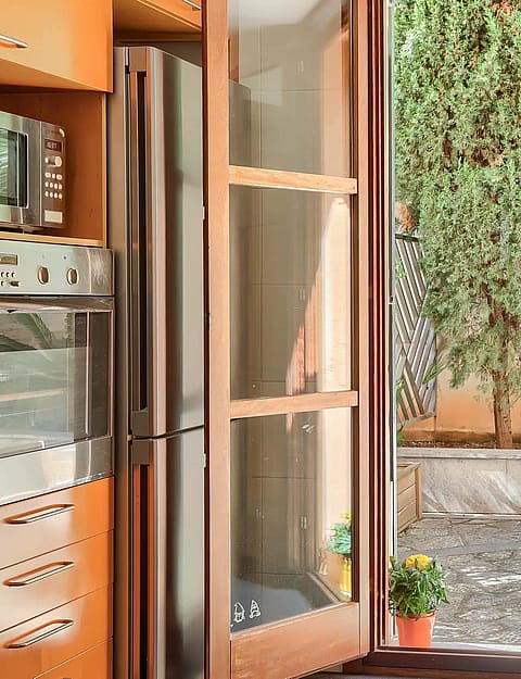 A kitchen interior featuring warm wooden cabinetry, a glass-topped table with fruit, and full-height glass doors leading out.