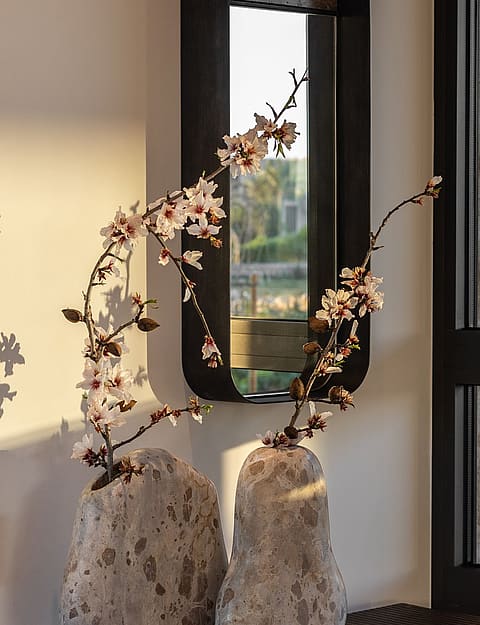 Close-up of flowering branches in stone vases on a surface near a stone wall and modern black-framed window.