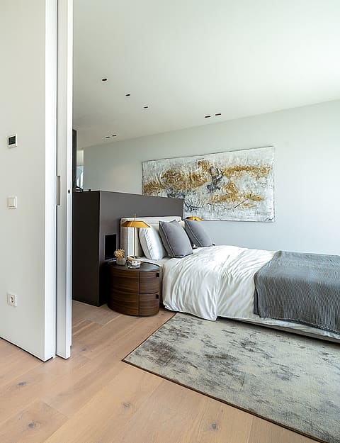 An open-plan bedroom with light wood floors and a glass-enclosed shower room, featuring modern fixtures and neutral tones.