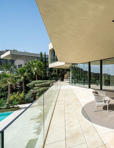 A wide, covered outdoor terrace with beige stone flooring, a modular sofa on a circular rug, and glass balustrades.