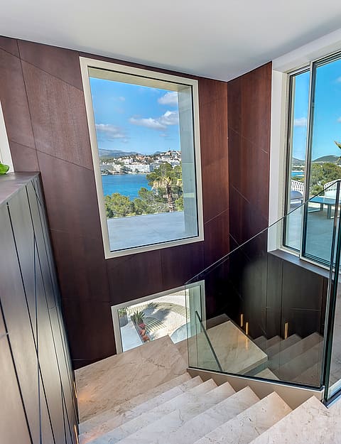 A high-angle view looking down a marble staircase with a dark wooden feature wall, glass balustrades, and large windows looking out toward a blue bay.