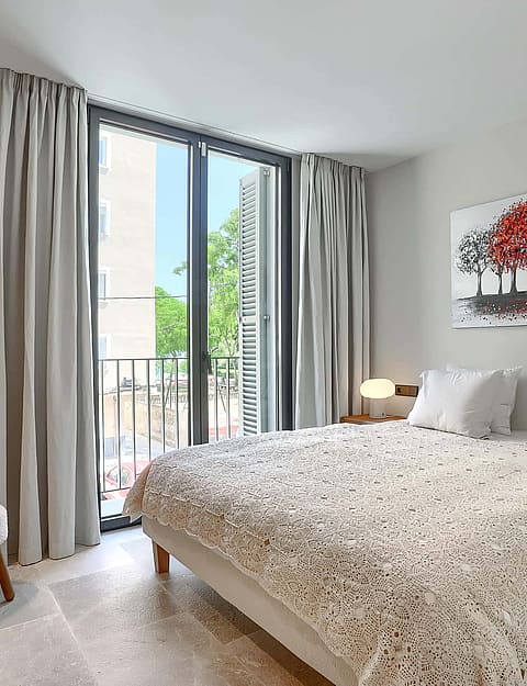 A contemporary bedroom featuring a queen bed with white bedding, a balcony door with grey curtains, and a textured armchair.