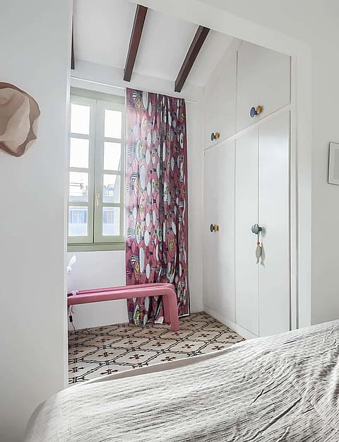 A bedroom featuring white built-in wardrobes, a patterned rug, and a pink bench near a window with floral curtains.