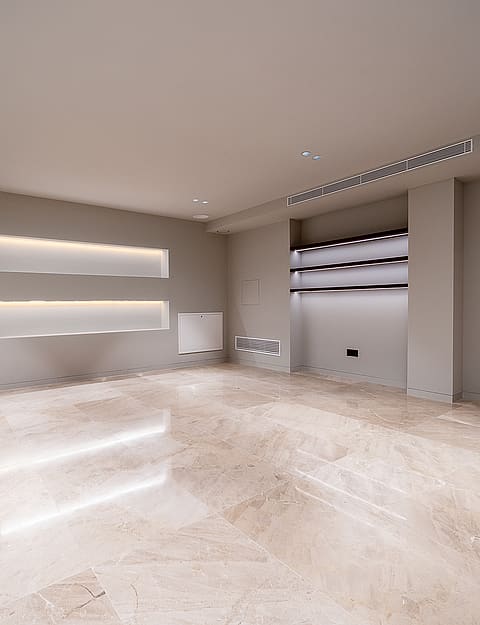 A spacious, unfurnished room with beige stone floors, recessed wall shelving with integrated LED lighting, and neutral walls.
