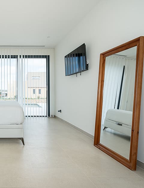 A minimalist bedroom featuring a white queen bed, light stone flooring, and a large wooden-framed floor mirror.