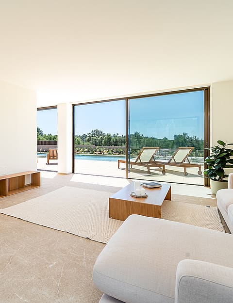 Bright open-plan living room featuring neutral sofas, a woven wood armchair, and floor-to-ceiling glass doors opening to a pool deck.