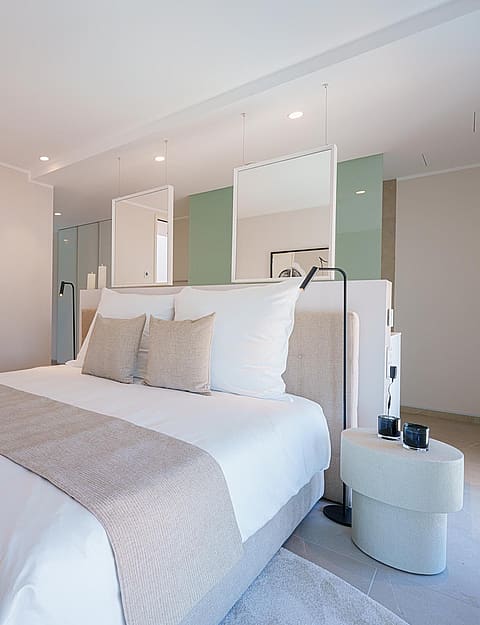 Dormitorio moderno con cama central, ropa de cama blanca y tonos beige sobre paredes de color claro.