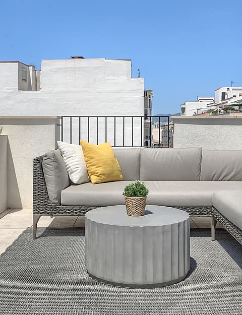 A rooftop terrace in El Molinar featuring a grey modular sofa with yellow cushions, a round fluted concrete table, and a patterned rug against a bright sky.