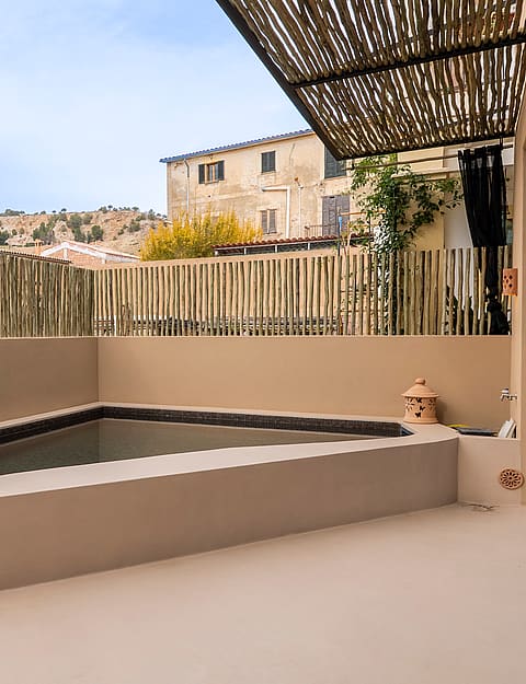 A small corner swimming pool on a terrace with beige stucco walls, bamboo fencing, and a reed shade cover.