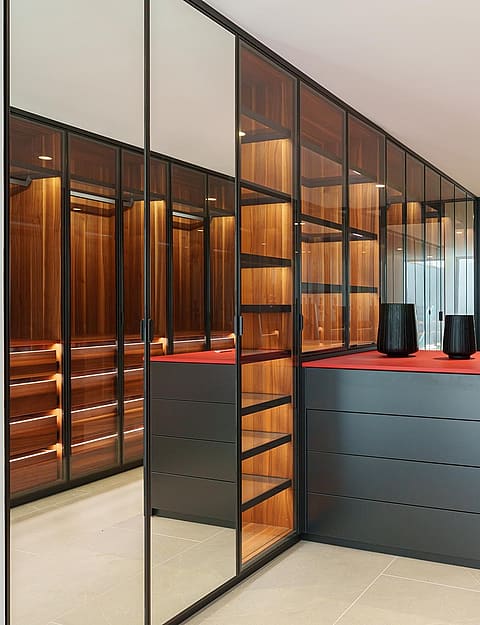 A walk-in dressing room featuring dark wood shelving enclosed by glass panels, with a central dark matte island unit on a light stone floor.