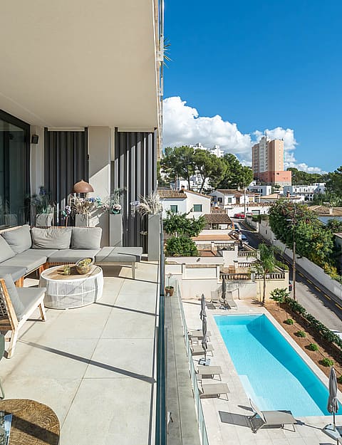 A covered terrace with grey sectional seating overlooks a rectangular swimming pool surrounded by a paved garden in a suburban setting.