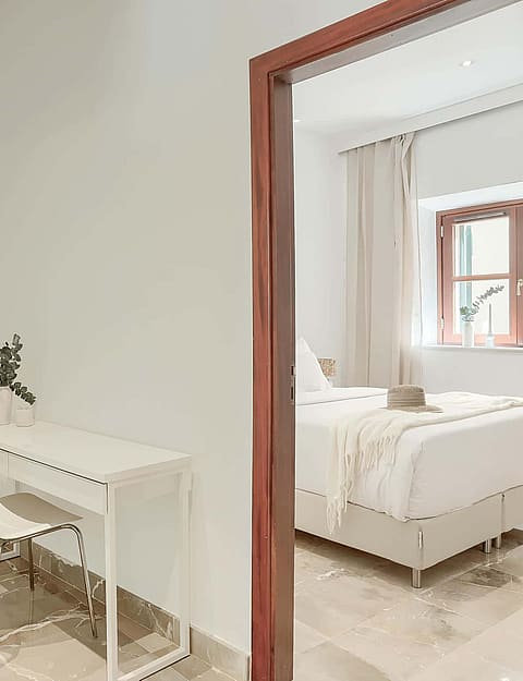 A small minimalist study area with a white console desk and chair, located in an alcove next to a doorway showing a bedroom.