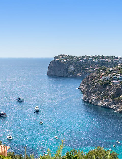 Elevated coastal view showing anchored yachts in a bright blue bay surrounded by rugged rocky cliffs and green hills.