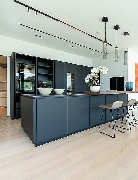 A modern kitchen with a dark grey central island, light timber flooring, and minimalist white cabinetry in a large open space.