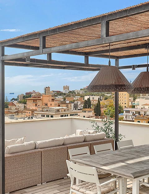 A rooftop terrace featuring a wicker sofa, dining set, and a pergola against a backdrop of the Mediterranean Sea and San Agustin.