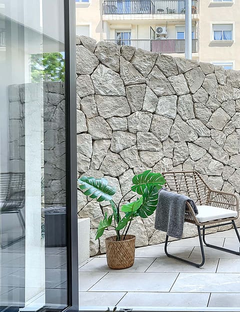 A private outdoor patio featuring a high dry-stone boundary wall, light-toned stone paving, and two rattan chairs with a small table.